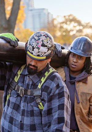 Man and woman with hard hats carrying large pipe together over their shoulder