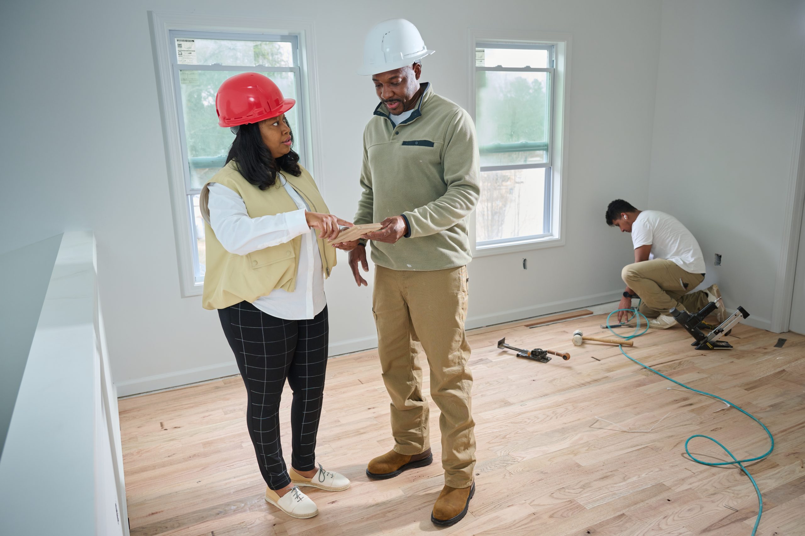 African American female and male in hard hats discussing construction plans in an interior room with a Hispanic male installing flooring in the background