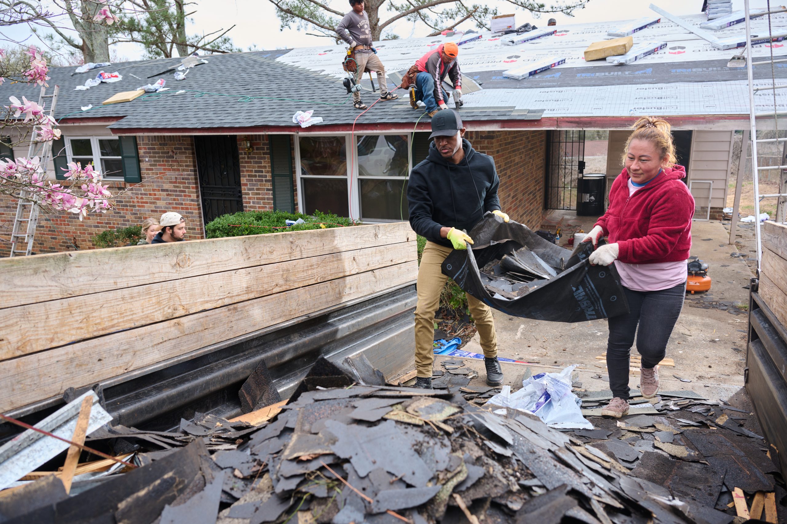 African American male and Hispanic female cleaning up a roofing job site with roofers working in the background